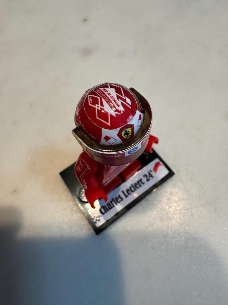 May include: A red and white Lego minifigure of a race car driver wearing a red helmet with a white Ferrari logo and the text "Scuderia Ferrari" on it. The minifigure is standing on a black base with the text "Charles Leclerc 24" on it.