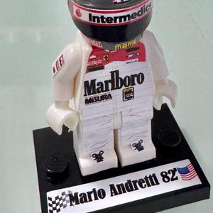 May include: A Lego figurine of Mario Andretti in a racing suit. The figure wears a helmet with "Intermedics" and "Marlboro" branding. The base features the name "Mario Andretti 82" with a checkered flag and American flag.