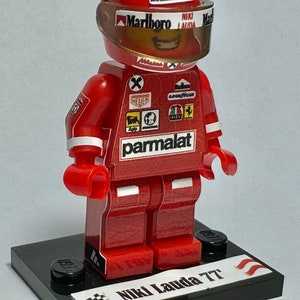 May include: A red Lego figurine of Niki Lauda, a Formula One racing driver, wearing a red racing suit, a helmet with a Marlboro logo, and a Parmalat sponsor logo. The figurine is standing on a black base with the text "Niki Lauda 77" and a chequered flag.