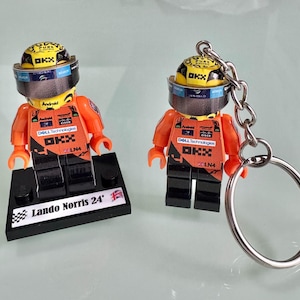 Può includere: Due minifigure Lego di piloti di corse che indossano tute da corsa arancioni e nere con caschi. La minifigure a sinistra è su una base nera con il testo "Lando Norris 24". La minifigure a destra è attaccata a un portachiavi argentato.