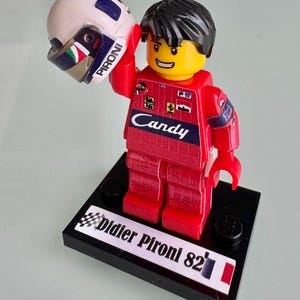 May include: A plastic minifigure wearing a red racing suit with Candy branding, holding a white helmet with Pironi text. The figure stands on a black base with a label reading Didier Pironi 82, featuring a checkered flag and a French flag icon.