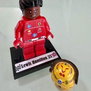May include: A plastic minifigure of a race car driver in a red racing suit with various sponsor logos, including HP and Shell. The figure stands on a black display base labeled Lewis Hamilton 25 with a British flag. A yellow racing helmet is nearby.
