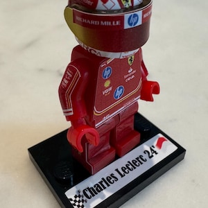May include: A red and white Lego minifigure of a race car driver wearing a red helmet with a white and red design. The figure is standing on a black base with a white sticker that says "Charles Leclerc 24".