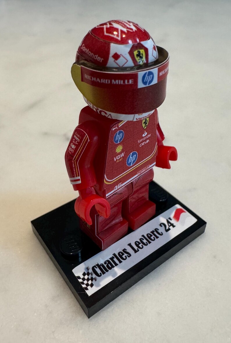 May include: A red and white Lego minifigure of a race car driver wearing a red helmet with a white and red design. The figure is standing on a black base with a white sticker that says "Charles Leclerc 24".