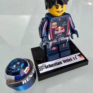 May include: A LEGO minifigure of a race car driver wearing a dark blue Red Bull racing suit with sponsor logos, standing on a black display base labeled Sebastian Vettel 11 with a German flag. A matching blue and white racing helmet sits nearby.