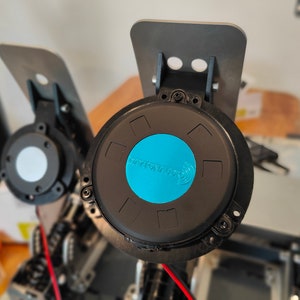 May include: Close-up of a black and blue round pedal with the text "DYNOAUTO" on the blue center. The pedal is attached to a black metal bracket.