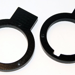 Op de afbeelding: Twee zwarte plastic ringen met een klein lipje aan één kant. De ringen hebben een ronde opening in het midden.