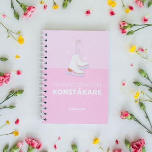 Op de afbeelding: Een roze spiraalgebonden notitieboek met de tekst "TRÄNINGSBOK FÖR KONSTÅKARE" en een illustratie van witte schaatsen. Het notitieboek is omgeven door roze en gele bloemen op een witte achtergrond.