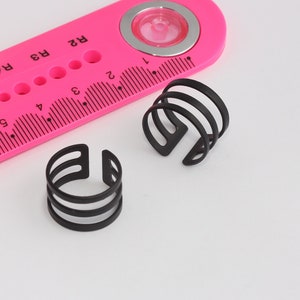 Puede incluir: Dos anillos de metal negro con un diseño de triple banda. Los anillos están sobre una superficie blanca junto a una regla rosa.