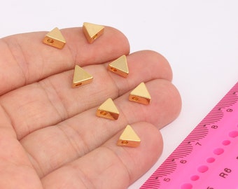 May include: A hand holding seven gold triangle-shaped metal beads. The beads are about 1 cm in size and have a hole in the center. A pink ruler is in the background.