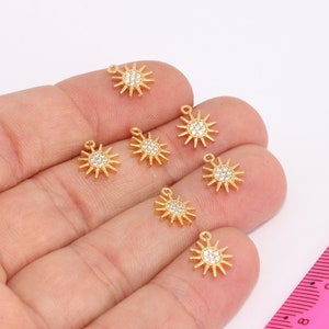 May include: A close-up of seven gold-toned sun charms with clear crystal accents. The charms are arranged on a hand and a pink ruler.