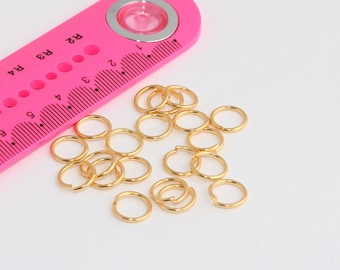 May include: A pink ruler with a circle in the center and a scale marked in inches and fractions of an inch. The ruler is next to a pile of gold jump rings.
