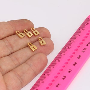 Puede incluir: Cinco pequeños encantos de candado de color dorado. Los encantos están dispuestos en una mano, con una regla rosa en el fondo.