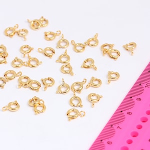 Può includere: Primo piano di un gruppo di 30 chiusure a moschettone in metallo dorato sparse su una superficie bianca. Un righello rosa è visibile nell'angolo in basso a destra dell'immagine.