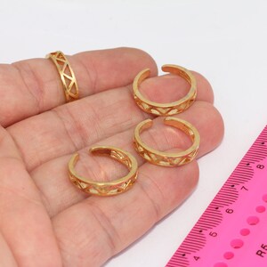 May include: Four gold toe rings with a geometric design. The rings are on a hand with a pink ruler in the background.