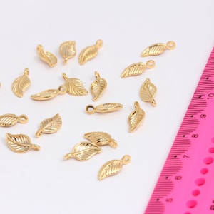 May include: A collection of 17 gold leaf charms with a loop at the top. The charms are scattered on a white surface with a pink ruler in the background.