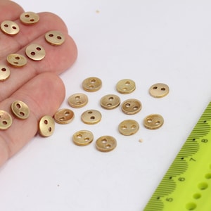 Puede incluir: Primer plano de una colección de botones de metal dorado con dos agujeros cada uno. Los botones están esparcidos sobre una superficie blanca, con algunos botones en una mano.