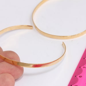 Pode incluir: Dois braceletes de metal dourado, um é ligeiramente maior que o outro, deitados sobre uma superfície branca ao lado de uma régua rosa.