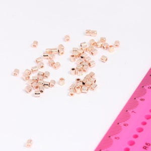Pode incluir: Um close-up de um monte de contas de metal cor de ouro rosa. As contas são pequenas e redondas, e estão espalhadas sobre uma superfície branca. Uma régua rosa é visível no fundo.