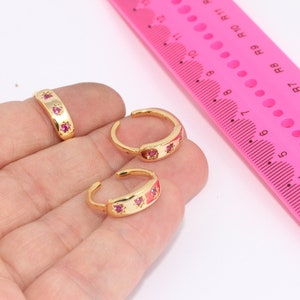 Könnte beinhalten: Drei goldene Ringe mit rosafarbenen Edelsteinen. Die Ringe befinden sich auf einer Hand und einem rosa Lineal mit Zahlen und Markierungen.
