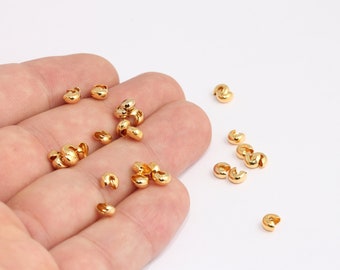 May include: A close-up of a hand holding a pile of gold-colored metal crimp beads. The beads are small and round, with a small opening on one side. The beads are scattered on a white surface.