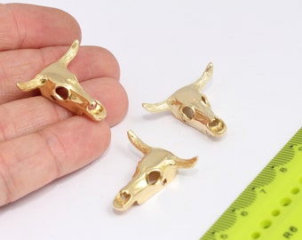 May include: Three gold-colored bull skull charms. Each charm features a detailed skull with horns and hollow eye sockets. The charms are displayed on a white surface next to a yellow ruler, suggesting a craft or jewelry-making theme.