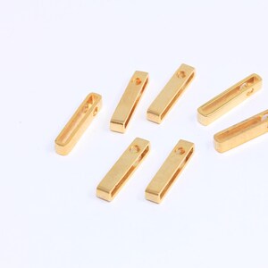 May include: Eight gold-colored metal rectangular jump rings with a small hole at the top. The rings are scattered on a white surface.