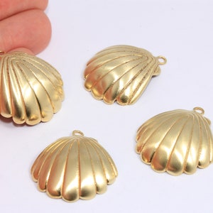 May include: Four gold-colored seashell charms with a textured, ridged design. Each charm has a small loop at the top for attaching to jewelry or other crafts. The charms are arranged on a white surface.
