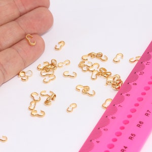 Può includere: Primo piano di un mucchio di anelli di salto in metallo dorato. Gli anelli sono sparsi su una superficie bianca accanto a un righello rosa con segni neri.
