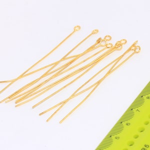 Può includere: Un gruppo di 10 spilli per la testa dorati con anelli a un'estremità. Gli spilli sono lunghi circa 5 cm e sono disposti in modo casuale su una superficie bianca.