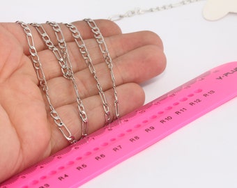 May include: A silver chain necklace with a figaro link pattern, laid out on a pink ruler with markings for ring sizes. The ruler is labeled "V-PLUS".