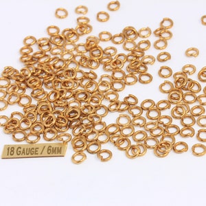 Op de afbeelding: Een grote stapel gouden springringen, 18 gauge en 6 mm groot. De ringen zijn verspreid over een wit oppervlak.  "18 Gauge / 6mm" is gedrukt op een gouden metalen label.