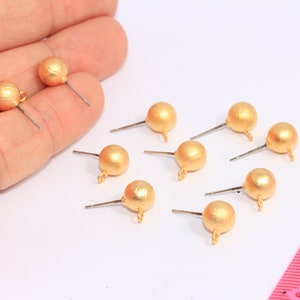 May include: Eight gold-toned earring findings with a round ball shape and a loop at the top. The findings are on a white surface with a pink ruler in the background.