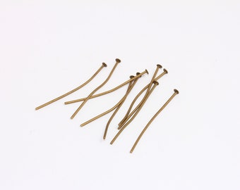 Può includere: Un gruppo di spilli per la testa dorati, noti anche come spilli per la creazione di gioielli, sono sparsi su una superficie bianca. Gli spilli sono lunghi circa 5 cm e hanno una piccola testa piatta.