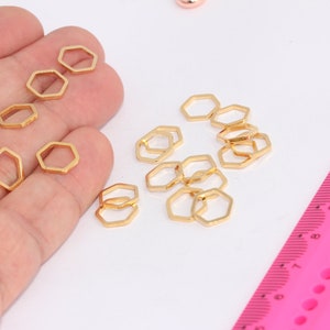 Könnte beinhalten: Nahaufnahme einer Hand, die eine Gruppe goldfarbener sechseckiger Sprungringe hält. Die Ringe sind auf einer weißen Oberfläche neben einem rosa Lineal verstreut.