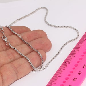 Può includere: Una collana a catena d'argento con una chiusura a moschettone. La catena è appoggiata su una mano e un righello rosa con segni in pollici.