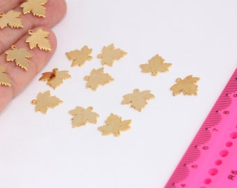 May include: A collection of 13 gold-toned metal maple leaf charms, scattered on a white surface. A pink ruler is visible in the background.