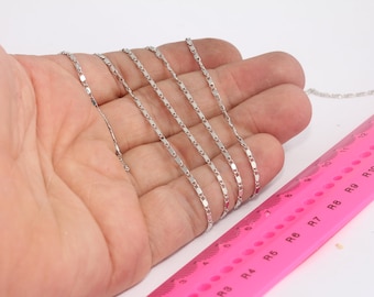 May include: Five silver chains of different lengths laid out on a hand, with a pink ruler in the background. The ruler is marked with numbers and the letters R and 8.