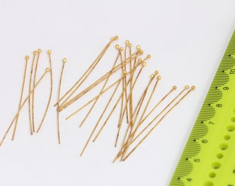 May include: A group of gold head pins with round heads. The pins are scattered on a white surface. A green ruler is in the background.