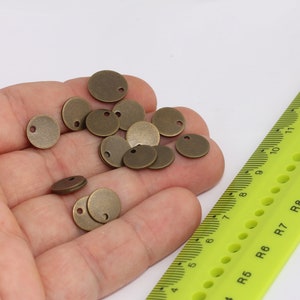 以下が含まれることがあります： 手の中に、中心に穴の開いた小さな丸いブロンズ色の金属製の円盤がいくつか置かれています。円盤はランダムなパターンで配置されています。背景には、黒い目盛りが付いた黄色の定規が見えます。