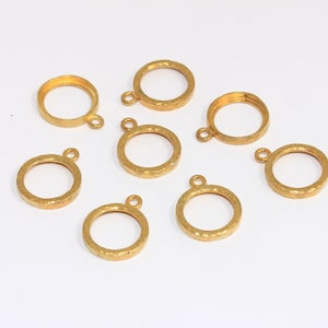 Puede incluir: Siete colgantes de anillo de metal dorado con una superficie texturizada y un pequeño lazo en la parte superior. Los colgantes son redondos y parecen ser para la fabricación de joyas o manualidades. Están dispuestos sobre un fondo blanco.