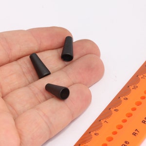 May include: Three black rubber cone-shaped fishing bobbers in a hand. A ruler with orange markings and black numbers is in the background.