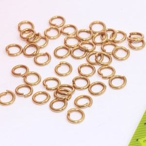 Op de afbeelding: Een stapel gouden springringen, een soort sieradenbevestiging, verspreid over een wit oppervlak. Een gele liniaal is zichtbaar op de achtergrond.