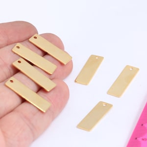 May include: Six gold-coloured metal rectangular tags with a hole at the top. The tags are arranged in a hand and on a pink ruler.