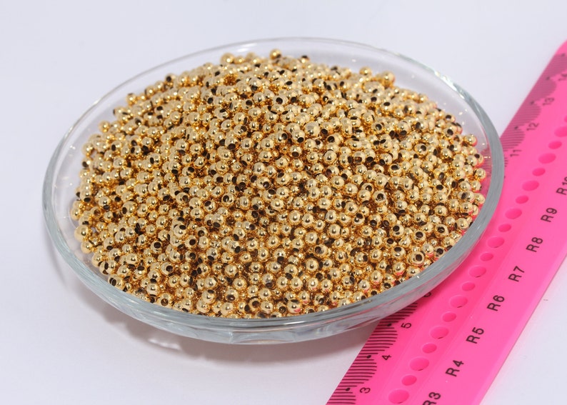 Op de afbeelding: Een glazen kom gevuld met een grote hoeveelheid kleine, glanzende goudkleurige metalen kralen. De kralen zijn rond en glad en zijn in een willekeurig patroon gerangschikt. Een roze liniaal met zwarte markeringen is zichtbaar op de achtergrond.