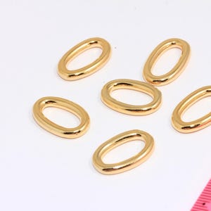 Könnte beinhalten: Sechs goldfarbene, ovale Schmuckanhänger liegen auf einer weißen Oberfläche. Ein rosa Lineal befindet sich in der unteren rechten Ecke. Die Anhänger haben eine glatte, polierte Oberfläche.