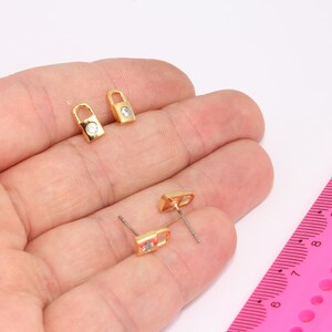 May include: A pair of gold padlock earrings with clear rhinestone accents. The earrings are shown on a hand against a white background.