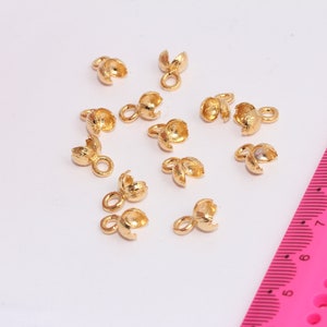 May include: A close-up of 12 gold-colored metal crimp beads with a loop on one end and two prongs on the other end. The beads are scattered on a white surface with a pink ruler in the background.