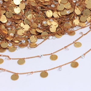Puede incluir: Tres cadenas doradas con pequeños colgantes redondos dorados y cuentas de cristal transparentes que cuelgan de ellas. Las cadenas están superpuestas y sobre un fondo blanco.