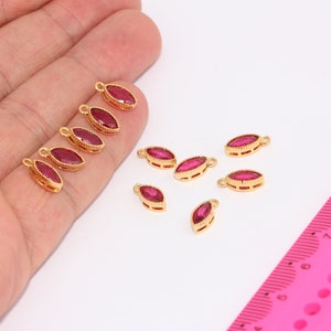 May include: A group of ten gold-toned marquise-shaped pendant charms with red crystal accents. The charms are displayed on a hand and a pink ruler.
